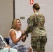 A female soldier in Army OCP (Operational Camouflage Pattern) uniform is presenting or pinning something to a smiling civilian woman seated in a chair. An older woman seated nearby observes the interaction with a smile. The setting appears to be an indoor military ceremony or event, likely a deployment, homecoming, or recognition ceremony.