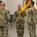 Four U.S. Army soldiers in OCP (Operational Camouflage Pattern) uniforms are participating in a military change of command or activation ceremony inside an indoor facility. A central soldier identified by a name tape reading 'JEFFERSON' is holding a unit guidon (ceremonial flag) with yellow fringe, passing or receiving it in what appears to be a formal military ceremony. The guidon features teal/green and yellow colors with what appears to be a unit crest.