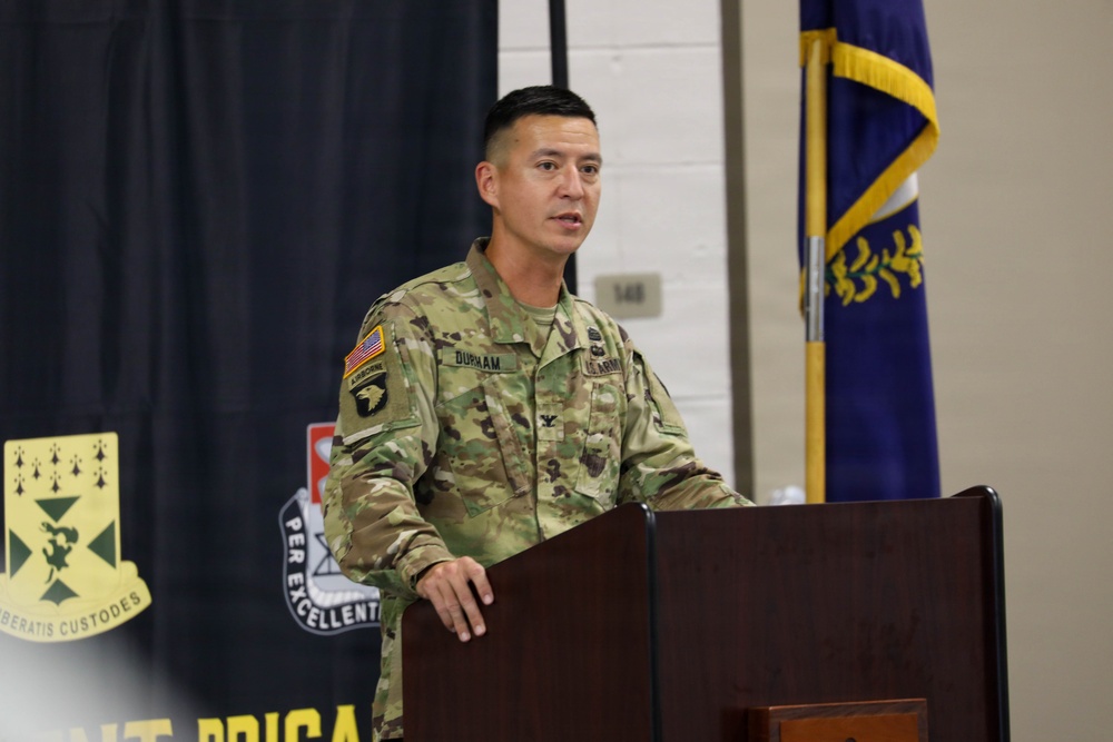 A U.S. Army Colonel named Durham is speaking at a podium during an official military ceremony or briefing. He is wearing the Army Combat Uniform (ACU) in MultiCam pattern with an Airborne tab and 101st Airborne Division patch visible on his left shoulder. The setting appears to be an indoor military facility with unit insignia banners and a military flag visible in the background.