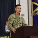 A U.S. Army Colonel named Durham is speaking at a podium during an official military ceremony or briefing. He is wearing the Army Combat Uniform (ACU) in MultiCam pattern with an Airborne tab and 101st Airborne Division patch visible on his left shoulder. The setting appears to be an indoor military facility with unit insignia banners and a military flag visible in the background.