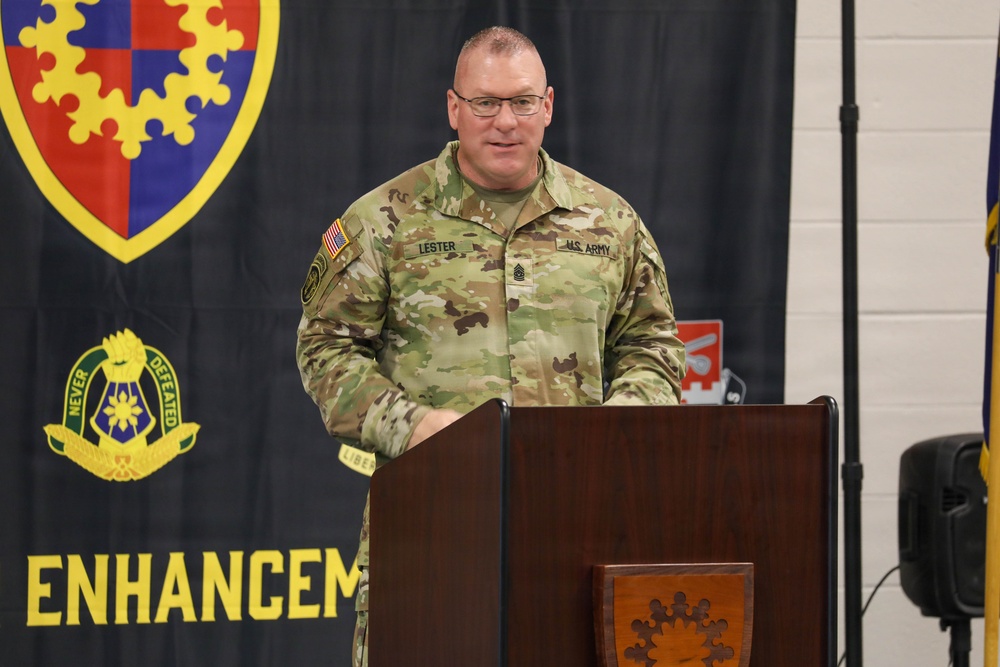 A U.S. Army soldier with the name tape 'LESTER' and senior NCO rank insignia is speaking at a wooden podium during an official military ceremony or event. He is wearing the Operational Camouflage Pattern (OCP) uniform with a U.S. flag patch and 'U.S. ARMY' tape visible. The backdrop features a military unit banner with a shield insignia, the motto 'NEVER DEFEATED,' and the partial text 'ENHANCEMENT.'