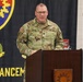 A U.S. Army soldier with the name tape 'LESTER' and senior NCO rank insignia is speaking at a wooden podium during an official military ceremony or event. He is wearing the Operational Camouflage Pattern (OCP) uniform with a U.S. flag patch and 'U.S. ARMY' tape visible. The backdrop features a military unit banner with a shield insignia, the motto 'NEVER DEFEATED,' and the partial text 'ENHANCEMENT.'