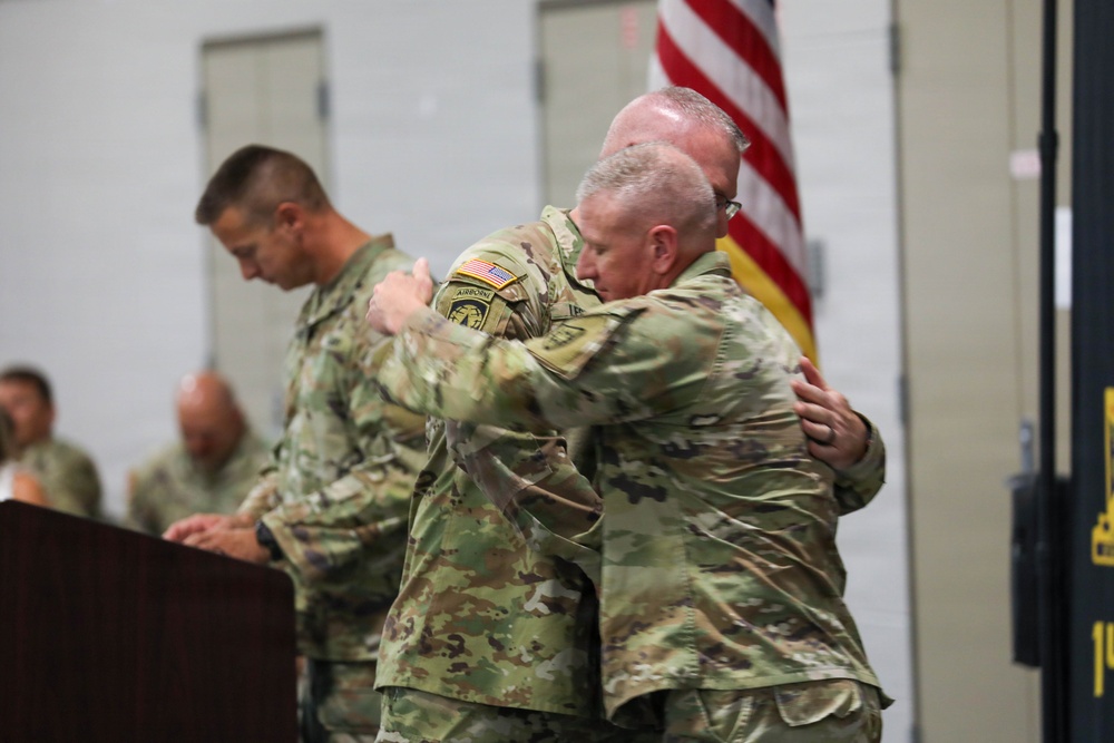 Two U.S. Army soldiers in OCP (Operational Camouflage Pattern) uniforms are embracing during what appears to be a military ceremony, likely a change of command or retirement event. An American flag is visible in the background, and additional uniformed personnel are present. One soldier has an Airborne tab visible on their left shoulder.