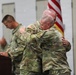 Two U.S. Army soldiers in OCP (Operational Camouflage Pattern) uniforms are embracing during what appears to be a military ceremony, likely a change of command or retirement event. An American flag is visible in the background, and additional uniformed personnel are present. One soldier has an Airborne tab visible on their left shoulder.