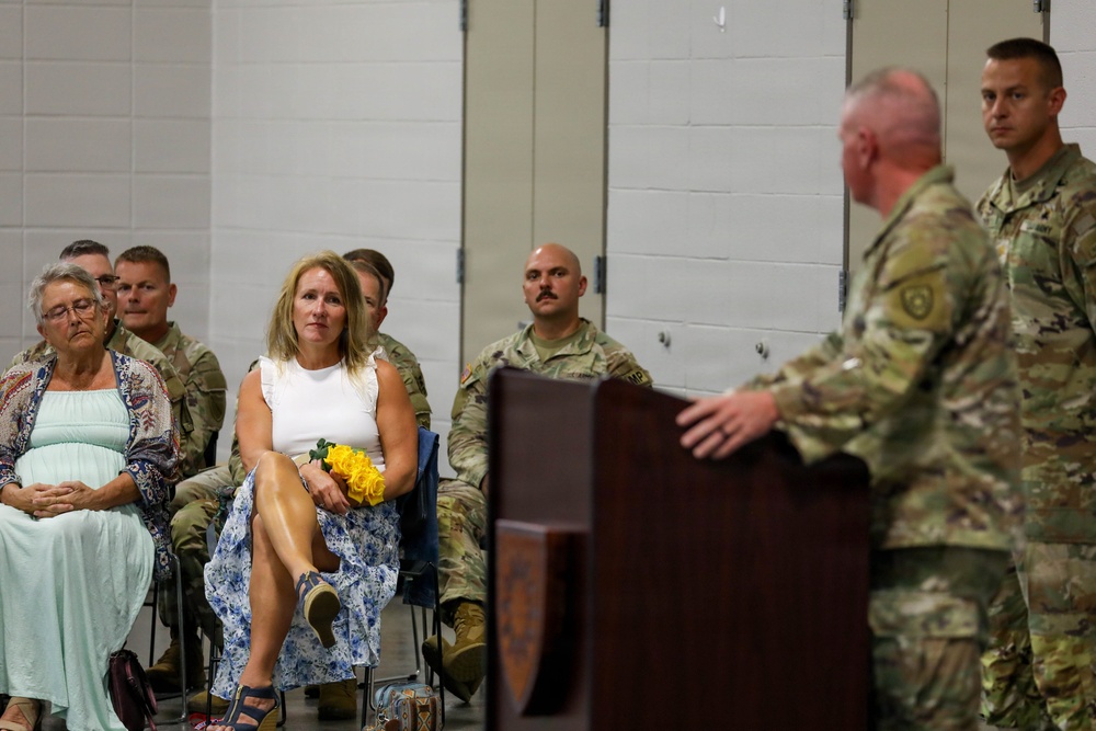 A military ceremony is taking place in what appears to be an indoor facility, likely a National Guard armory or military building. A uniformed soldier is speaking at a wooden podium while an audience of both military personnel in camouflage uniforms and civilian attendees listen attentively. A civilian woman in a white top and floral skirt holds yellow roses, suggesting she may be a family member being recognized during the ceremony.