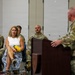 A military ceremony is taking place in what appears to be an indoor facility, likely a National Guard armory or military building. A uniformed soldier is speaking at a wooden podium while an audience of both military personnel in camouflage uniforms and civilian attendees listen attentively. A civilian woman in a white top and floral skirt holds yellow roses, suggesting she may be a family member being recognized during the ceremony.