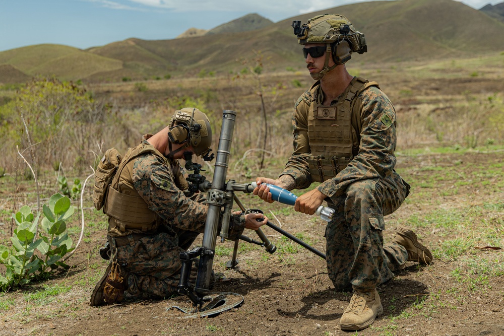 Two military personnel in MARPAT camouflage uniforms are operating what appears to be an M224 60mm mortar system in an open field training environment. One soldier is kneeling and adjusting the mortar tube while the other holds what appears to be a training round or inert munition. The setting appears to be a military training exercise based on the open terrain and mountainous background.