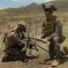 Two military personnel in MARPAT camouflage uniforms are operating what appears to be an M224 60mm mortar system in an open field training environment. One soldier is kneeling and adjusting the mortar tube while the other holds what appears to be a training round or inert munition. The setting appears to be a military training exercise based on the open terrain and mountainous background.