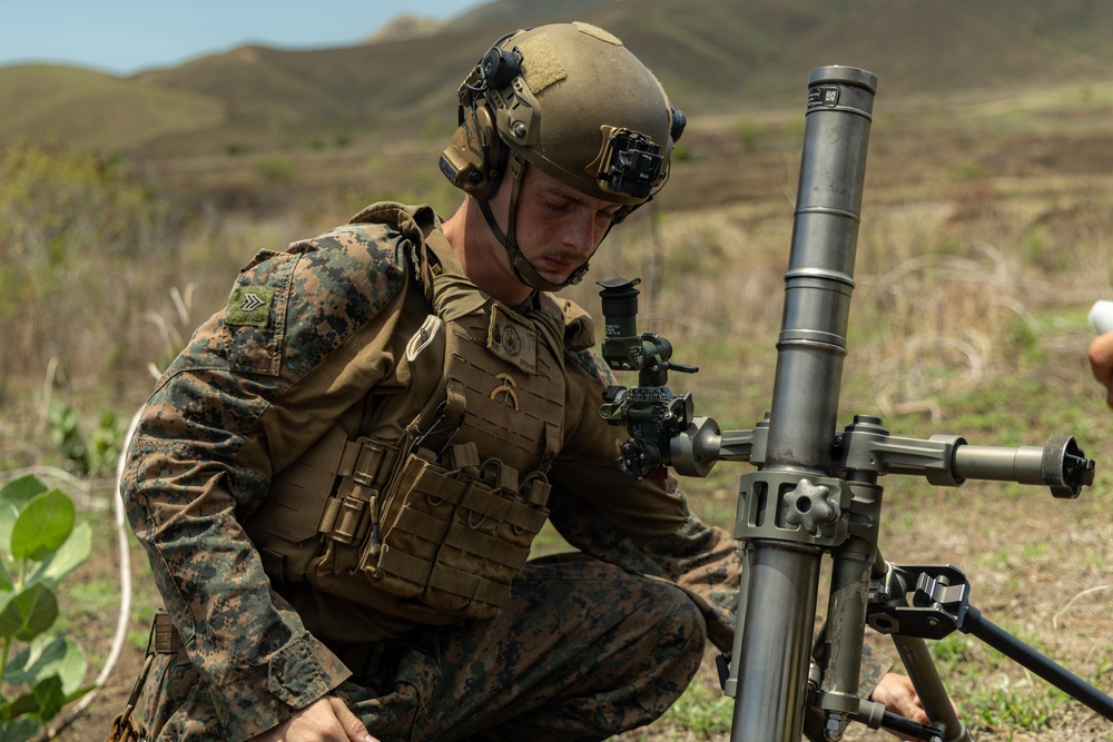 A U.S. Marine in full combat gear is kneeling and operating what appears to be an M224 60mm mortar system in an outdoor field environment. The soldier is wearing MARPAT digital camouflage, a tactical plate carrier, and a ballistic helmet with communications headset. The setting appears to be a training exercise based on the open terrain and visible hills in the background.