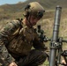 A U.S. Marine in full combat gear is kneeling and operating what appears to be an M224 60mm mortar system in an outdoor field environment. The soldier is wearing MARPAT digital camouflage, a tactical plate carrier, and a ballistic helmet with communications headset. The setting appears to be a training exercise based on the open terrain and visible hills in the background.