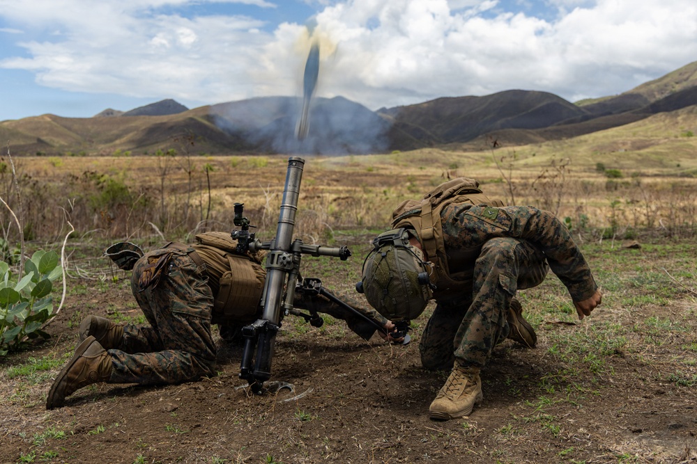 Two military personnel in camouflage uniforms are operating a mortar system in an open field, with a round visibly in flight and smoke rising in the background. The terrain features dry grassland with rolling hills and mountains in the distance. The image captures the moment immediately after a mortar has been fired.