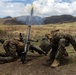Two military personnel in camouflage uniforms are operating a mortar system in an open field, with a round visibly in flight and smoke rising in the background. The terrain features dry grassland with rolling hills and mountains in the distance. The image captures the moment immediately after a mortar has been fired.