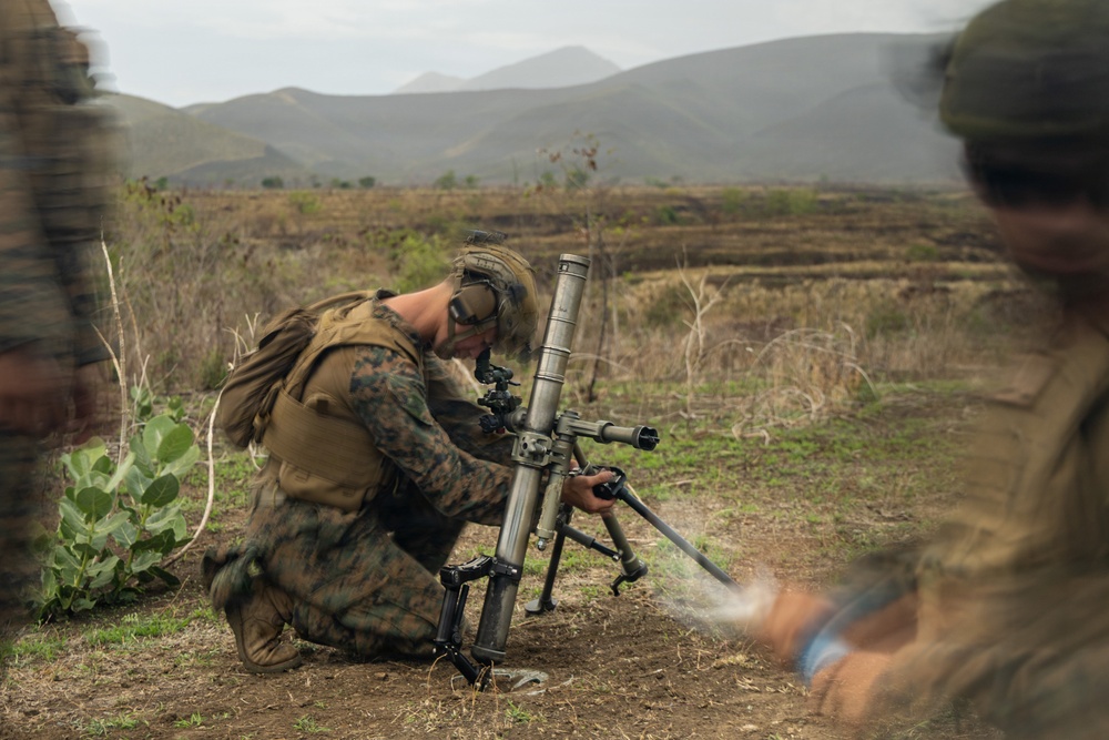 A soldier in digital camouflage uniform is kneeling on the ground operating a mortar system in an open field environment. The setting appears to be a training exercise or field operation in a semi-arid, hilly terrain. At least two additional personnel are partially visible at the edges of the frame.