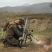 A soldier in digital camouflage uniform is kneeling on the ground operating a mortar system in an open field environment. The setting appears to be a training exercise or field operation in a semi-arid, hilly terrain. At least two additional personnel are partially visible at the edges of the frame.