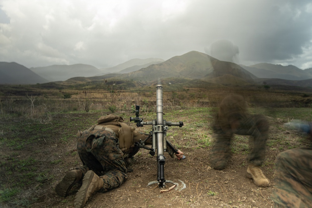 A military service member in camouflage uniform is operating a mortar system in an open field with mountainous terrain in the background. The image captures the moment of or immediately after firing, as evidenced by visible smoke/blast disturbance near the weapon. The overcast sky and rugged landscape suggest a training exercise environment.