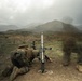 A military service member in camouflage uniform is operating a mortar system in an open field with mountainous terrain in the background. The image captures the moment of or immediately after firing, as evidenced by visible smoke/blast disturbance near the weapon. The overcast sky and rugged landscape suggest a training exercise environment.
