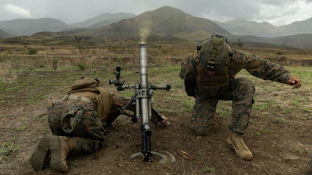 Two military personnel in digital camouflage uniforms are operating a mortar system in an open field. Smoke is visible rising from the mortar tube, indicating a round has just been fired. The setting appears to be a training exercise in a mountainous, overcast environment.