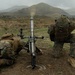 Two military personnel in digital camouflage uniforms are operating a mortar system in an open field. Smoke is visible rising from the mortar tube, indicating a round has just been fired. The setting appears to be a training exercise in a mountainous, overcast environment.