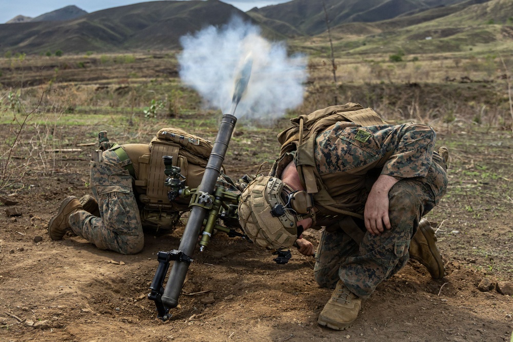 Two U.S. Marines in MARPAT camouflage uniforms are operating a mortar system in a field training environment. The mortar has just been fired, as evidenced by the visible smoke and propellant gases exiting the tube. The setting appears to be a hilly, semi-arid training range.