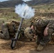 Two U.S. Marines in MARPAT camouflage uniforms are operating a mortar system in a field training environment. The mortar has just been fired, as evidenced by the visible smoke and propellant gases exiting the tube. The setting appears to be a hilly, semi-arid training range.