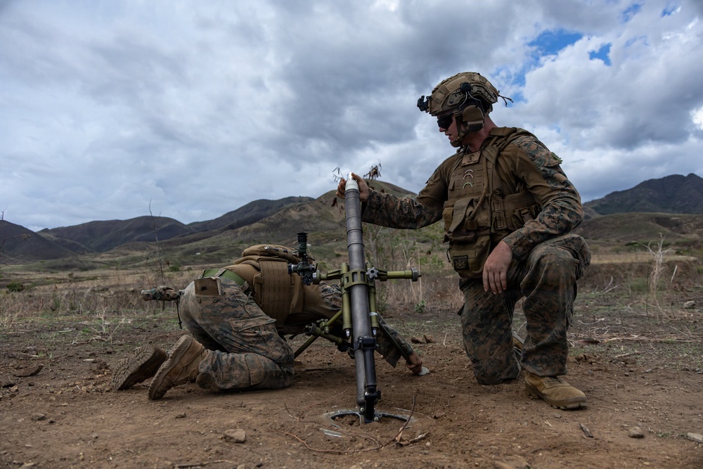 Two U.S. Marines in full combat gear are operating a mortar system in an open, arid training area with mountainous terrain in the background. One Marine is kneeling and preparing to drop a mortar round into the tube while the second Marine is positioned prone, likely observing or adjusting the sight. The setting and uniforms are consistent with a U.S. Marine Corps field training exercise.