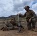 Two U.S. Marines in full combat gear are operating a mortar system in an open, arid training area with mountainous terrain in the background. One Marine is kneeling and preparing to drop a mortar round into the tube while the second Marine is positioned prone, likely observing or adjusting the sight. The setting and uniforms are consistent with a U.S. Marine Corps field training exercise.