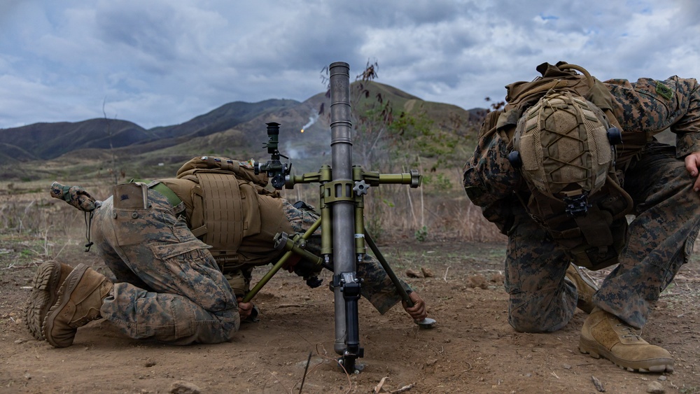Two military personnel in MARPAT (Marine pattern) camouflage uniforms are operating a mortar system in a field training environment. A projectile or smoke trail is visible in the background sky, indicating the mortar has just been fired. The setting appears to be a mountainous training range with overcast skies.
