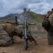 Two military personnel in MARPAT (Marine pattern) camouflage uniforms are operating a mortar system in a field training environment. A projectile or smoke trail is visible in the background sky, indicating the mortar has just been fired. The setting appears to be a mountainous training range with overcast skies.