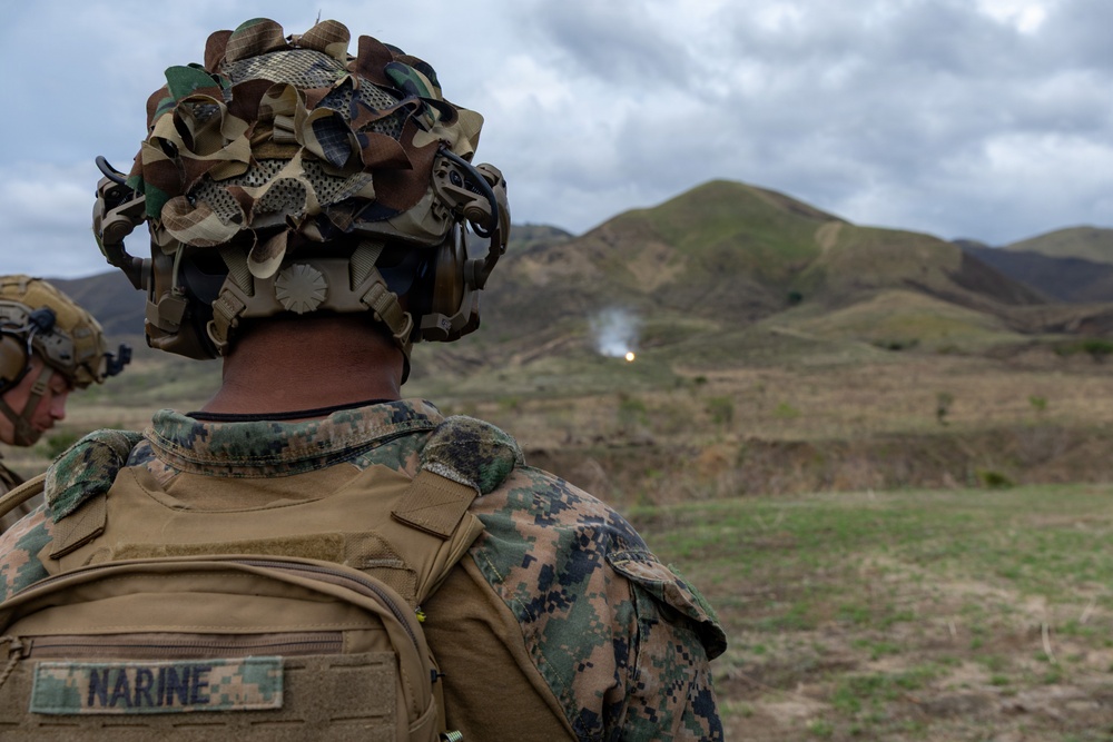 A U.S. Marine identified by the name tape 'NARINE' on their body armor observes a live-fire exercise from a training range. The Marine is wearing MARPAT digital camouflage, a camouflage-covered helmet with a ghillie-style cover, and tactical hearing protection. In the background, an explosion or impact flash with smoke is visible against a hilly, semi-arid terrain.