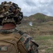 A U.S. Marine identified by the name tape 'NARINE' on their body armor observes a live-fire exercise from a training range. The Marine is wearing MARPAT digital camouflage, a camouflage-covered helmet with a ghillie-style cover, and tactical hearing protection. In the background, an explosion or impact flash with smoke is visible against a hilly, semi-arid terrain.