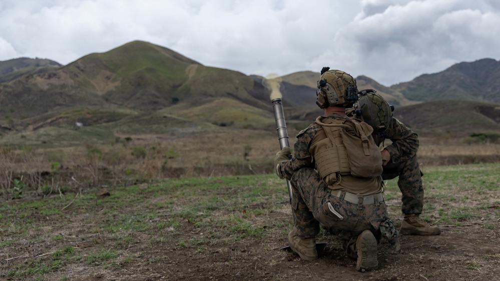 Two military personnel in full combat gear are positioned in a kneeling stance in an open field with mountainous terrain in the background. A projectile or rocket can be seen in flight, suggesting a weapon has just been fired. The soldiers are wearing MARPAT (Marine pattern) camouflage uniforms, consistent with U.S. Marine Corps equipment.