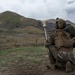 Two military personnel in full combat gear are positioned in a kneeling stance in an open field with mountainous terrain in the background. A projectile or rocket can be seen in flight, suggesting a weapon has just been fired. The soldiers are wearing MARPAT (Marine pattern) camouflage uniforms, consistent with U.S. Marine Corps equipment.