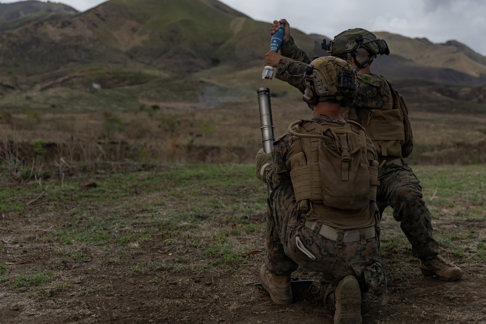 Two military personnel in full combat gear are operating a mortar system in an open field with mountainous terrain in the background. One soldier is kneeling and holding the mortar tube while the other is preparing to drop a round into the tube. The setting appears to be a training exercise based on the controlled environment and visible safety procedures being followed.