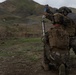 Two military personnel in full combat gear are operating a mortar system in an open field with mountainous terrain in the background. One soldier is kneeling and holding the mortar tube while the other is preparing to drop a round into the tube. The setting appears to be a training exercise based on the controlled environment and visible safety procedures being followed.