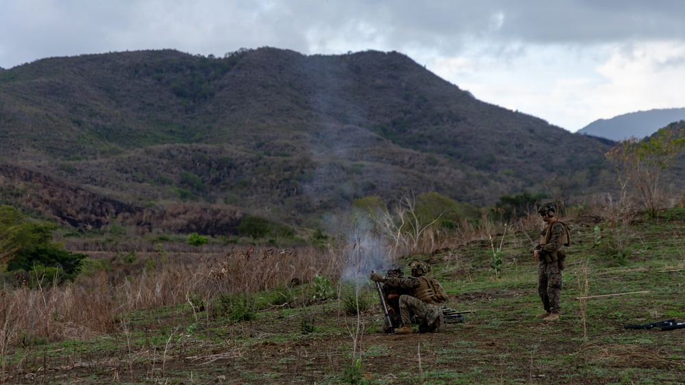 Military personnel in camouflage uniforms are operating what appears to be a mortar system in an open field, with visible smoke rising from the weapon. The scene takes place in a hilly, semi-arid landscape with sparse vegetation and overcast skies. At least three soldiers are present, with two crouched near the weapon system and one standing nearby observing.