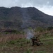 Military personnel in camouflage uniforms are operating what appears to be a mortar system in an open field, with visible smoke rising from the weapon. The scene takes place in a hilly, semi-arid landscape with sparse vegetation and overcast skies. At least three soldiers are present, with two crouched near the weapon system and one standing nearby observing.
