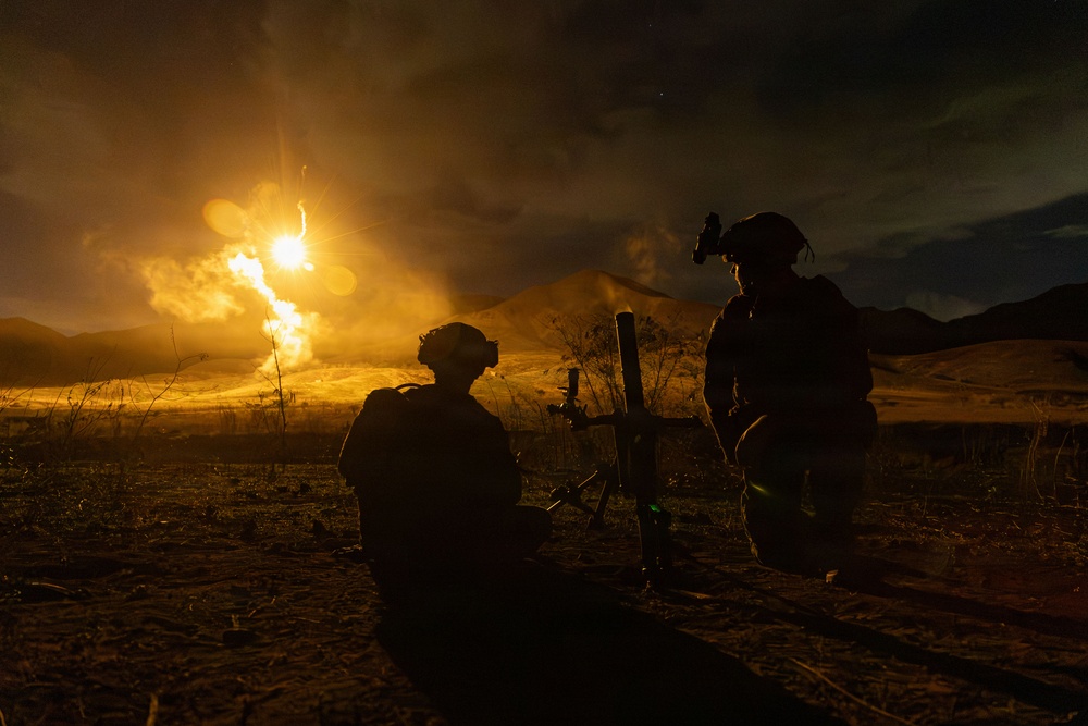 A nighttime military training or combat operation showing two soldiers in silhouette operating what appears to be a mortar system. A large explosion or illumination round is visible in the background, casting intense orange light across a desert or arid terrain with hills. The dramatic lighting contrast between the bright blast and dark surroundings creates a high-contrast tactical scene.
