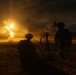 A nighttime military training or combat operation showing two soldiers in silhouette operating what appears to be a mortar system. A large explosion or illumination round is visible in the background, casting intense orange light across a desert or arid terrain with hills. The dramatic lighting contrast between the bright blast and dark surroundings creates a high-contrast tactical scene.