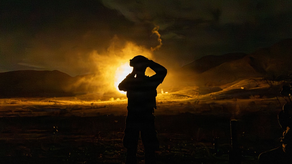 A silhouetted figure, appearing to be a military or tactical personnel, stands in the foreground observing a large explosion or controlled detonation in a desert/mountainous landscape at night. The bright orange and yellow fireball illuminates the surrounding terrain and smoke cloud from behind the figure. The dramatic backlighting creates a stark silhouette effect against the intense light of the blast.
