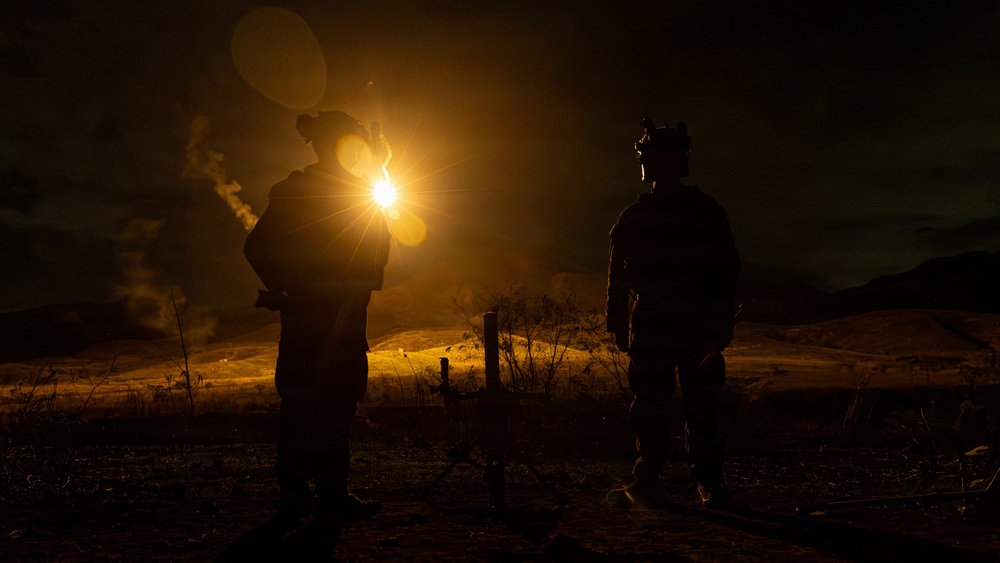 A nighttime outdoor scene showing two silhouetted figures standing in an open field or rural terrain. One figure appears to be holding a bright light source that creates a strong lens flare effect, illuminating the surrounding landscape in warm golden tones. Both figures appear to be wearing tactical or military-style gear including helmets.