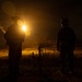 A nighttime outdoor scene showing two silhouetted figures standing in an open field or rural terrain. One figure appears to be holding a bright light source that creates a strong lens flare effect, illuminating the surrounding landscape in warm golden tones. Both figures appear to be wearing tactical or military-style gear including helmets.