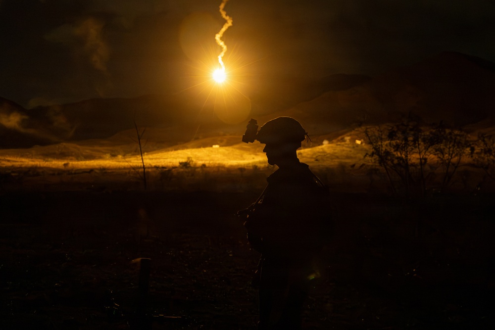 A silhouetted military personnel is visible in a nighttime outdoor setting, illuminated by a bright aerial illumination flare descending in the background. The flare casts a warm golden-orange light across a hilly, open terrain. The scene appears to depict a nighttime military training exercise or field operation.