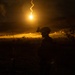 A silhouetted military personnel is visible in a nighttime outdoor setting, illuminated by a bright aerial illumination flare descending in the background. The flare casts a warm golden-orange light across a hilly, open terrain. The scene appears to depict a nighttime military training exercise or field operation.
