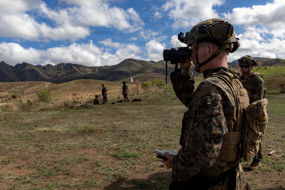 Military personnel in full combat gear are conducting field training or reconnaissance operations in a hilly, semi-arid outdoor environment. The foreground soldier is using binoculars while holding what appears to be a notepad or map, suggesting forward observation or fire support coordination. Additional personnel are visible in the background at varying distances across the terrain.