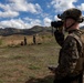 Military personnel in full combat gear are conducting field training or reconnaissance operations in a hilly, semi-arid outdoor environment. The foreground soldier is using binoculars while holding what appears to be a notepad or map, suggesting forward observation or fire support coordination. Additional personnel are visible in the background at varying distances across the terrain.