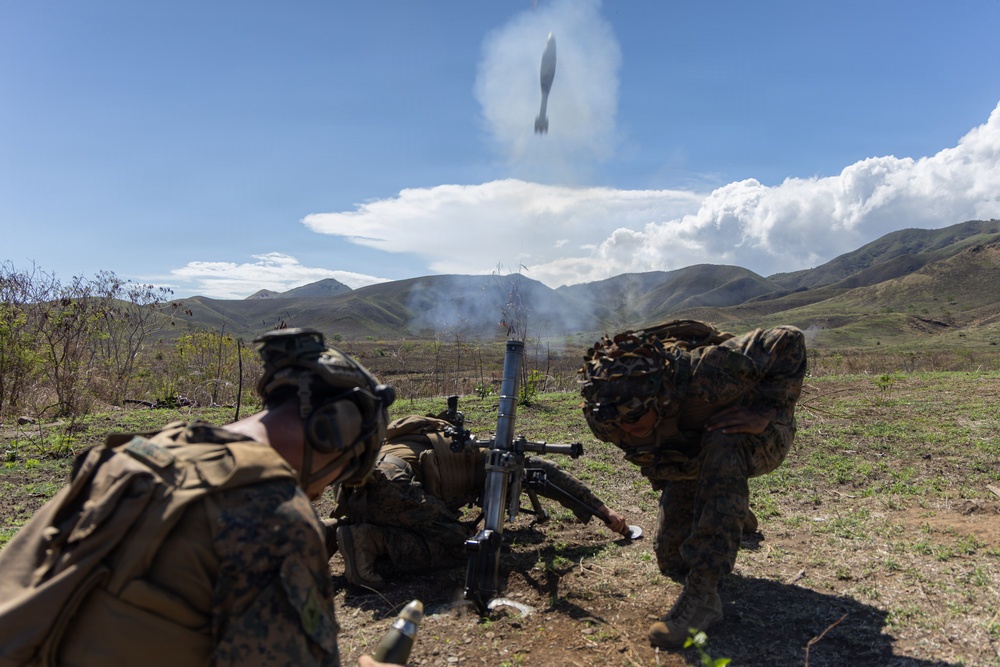 Military personnel in camouflage uniforms are operating a mortar system in an open field with mountainous terrain in the background. A mortar round is visibly captured mid-air just above the tube immediately after firing, with propellant smoke still rising from the weapon. The scene appears to be a live-fire training exercise based on the organized positioning of the soldiers.
