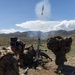 Military personnel in camouflage uniforms are operating a mortar system in an open field with mountainous terrain in the background. A mortar round is visibly captured mid-air just above the tube immediately after firing, with propellant smoke still rising from the weapon. The scene appears to be a live-fire training exercise based on the organized positioning of the soldiers.
