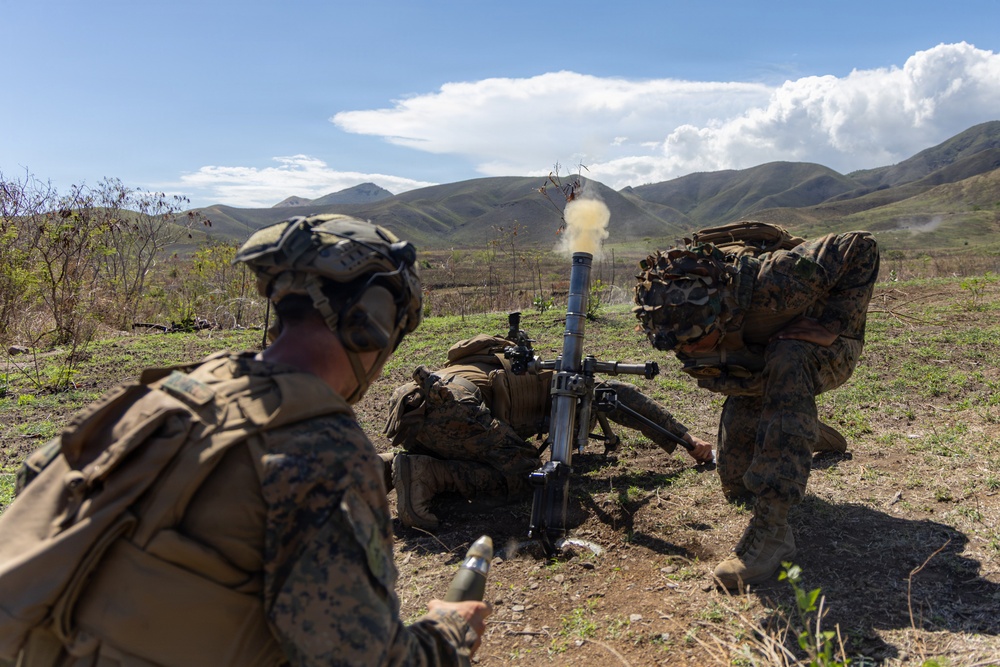 Military personnel in camouflage uniforms are operating a mortar system in an open field with mountainous terrain in the background. A round has just been fired, as evidenced by the smoke and propellant gases visible at the muzzle of the mortar tube. At least three soldiers are visible, each performing distinct roles in the mortar crew.