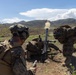 Military personnel in camouflage uniforms are operating a mortar system in an open field with mountainous terrain in the background. A round has just been fired, as evidenced by the smoke and propellant gases visible at the muzzle of the mortar tube. At least three soldiers are visible, each performing distinct roles in the mortar crew.