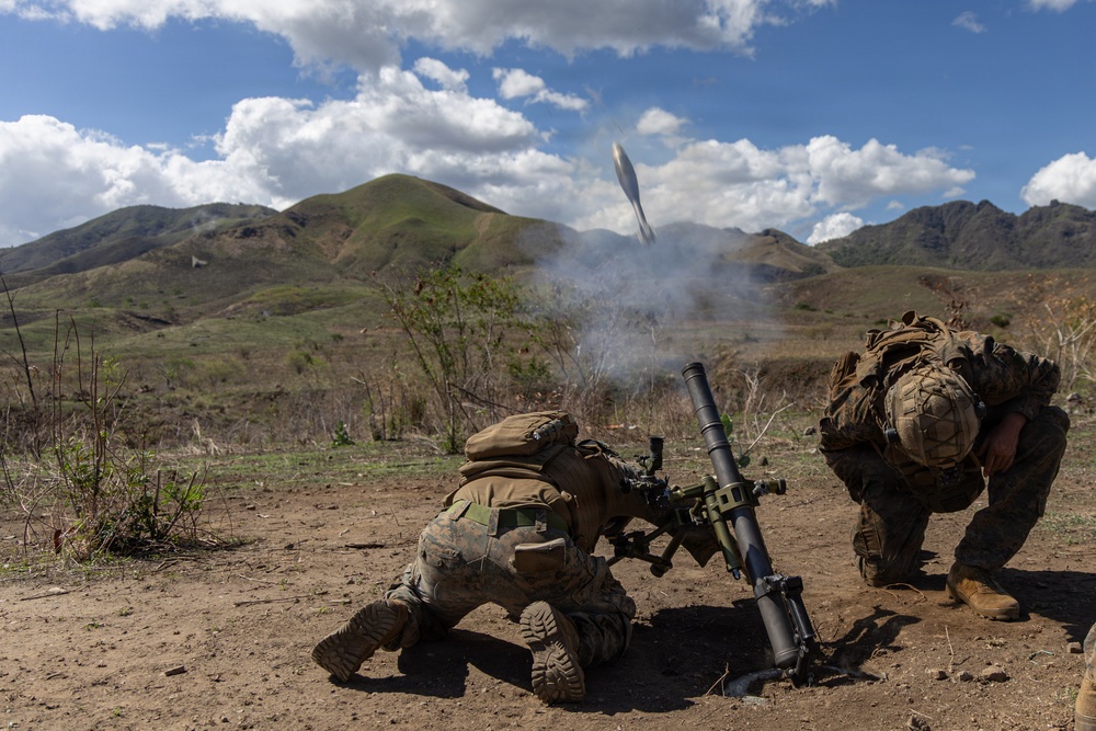 Two military personnel in full combat gear are operating a mortar system, with a round visibly in flight just after firing. The scene takes place in an open, arid training or operational area with green hills and mountains in the background. The image captures the moment of discharge with visible propellant smoke rising from the mortar tube.